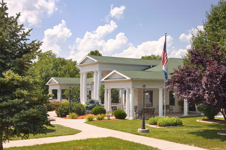 Exterior view of Canterbury Center showing a white building with green roofs, white columns, and a well-maintained lawn with trees and shrubs. There is a flagpole with an American flag and another flag in front of the building under a partly cloudy sky.