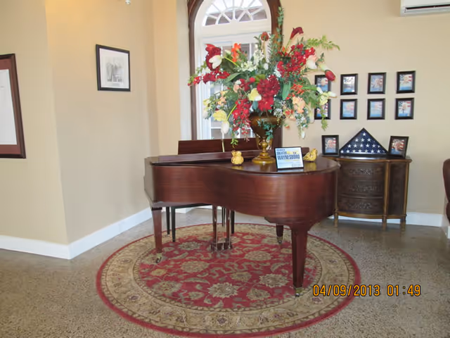Interior view of a room featuring a grand piano with a large floral arrangement on top, placed on a round decorative rug. Behind the piano, there is a small wooden cabinet with framed pictures and a folded American flag displayed on it. The walls are light-colored with framed pictures hanging, and a window with an arched top is visible in the background.