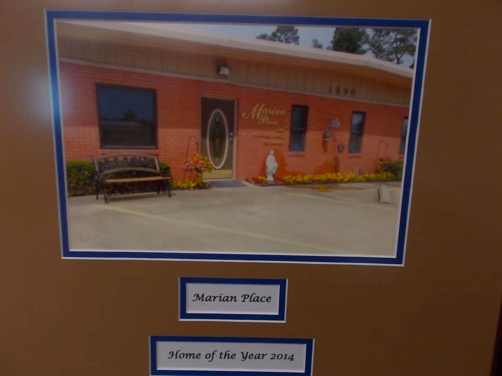 Framed photograph showing the front entrance of Marian Place with a bench and parking area, plus labels reading 'Marian Place' and 'Home of the Year 2014'.