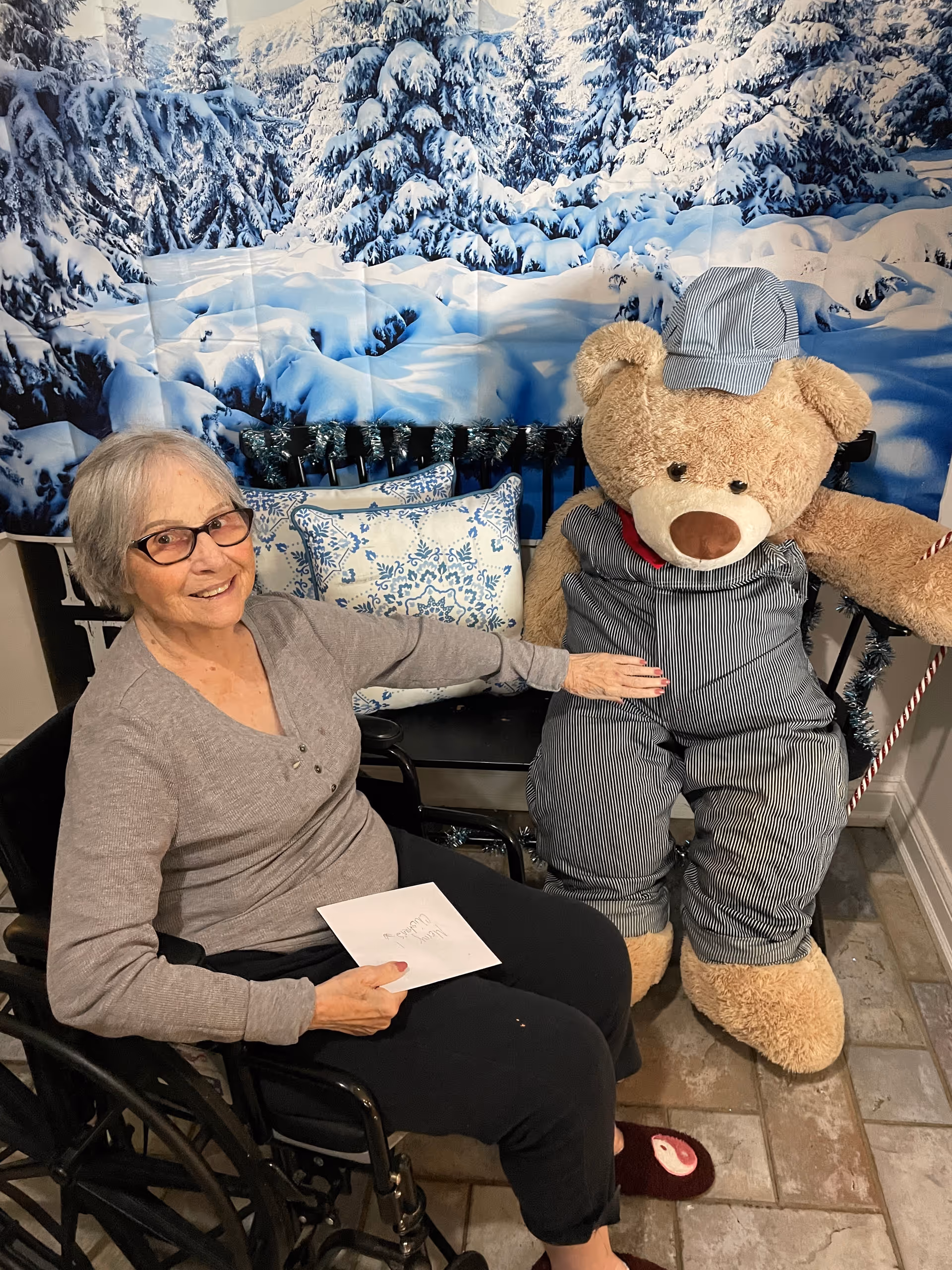 An elderly woman sitting in a wheelchair smiling and holding a card, next to a large teddy bear dressed in striped overalls and a matching hat. Behind them is a bench with decorative blue and white pillows and a backdrop featuring snow-covered trees.