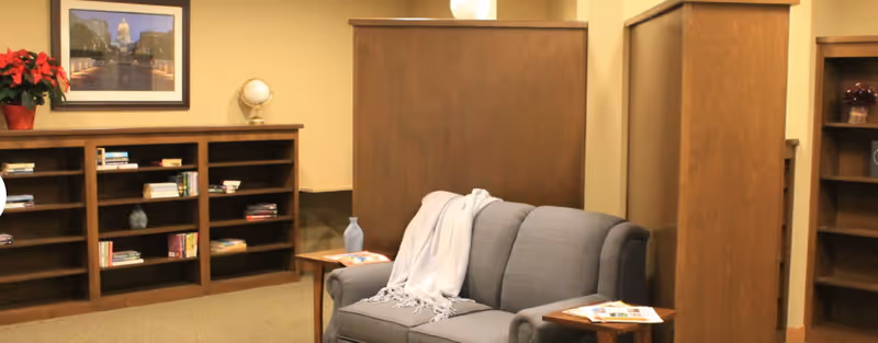 A cozy interior room with a gray upholstered sofa draped with a light-colored throw blanket, two wooden side tables with magazines, wooden bookshelves filled with books and decorative items, a framed picture on the wall, and a small globe on one of the shelves.