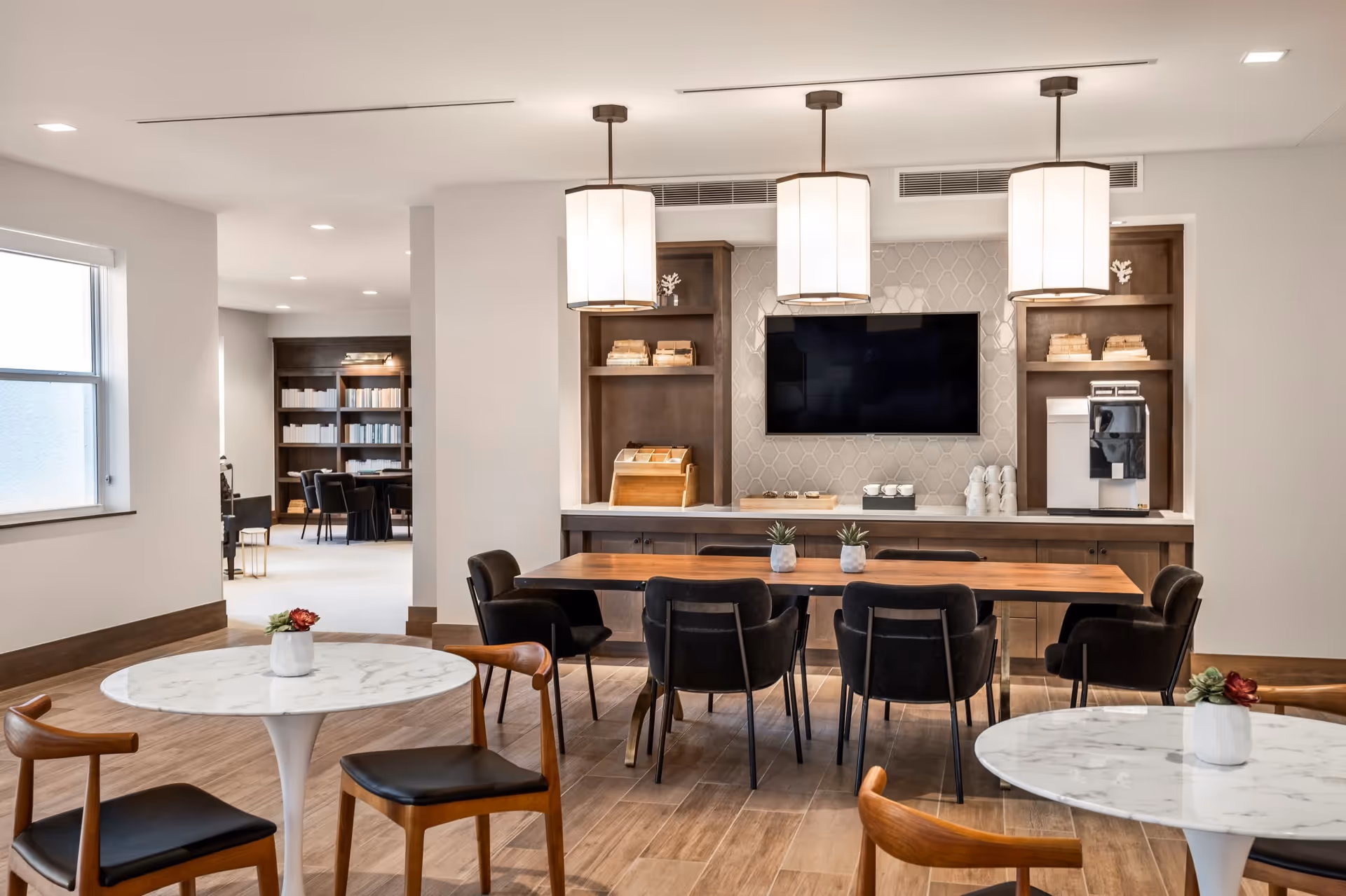 Bright modern communal dining and lounge area with marble-topped round tables, a long wooden table and chairs, a wall-mounted TV, and pendant lights.