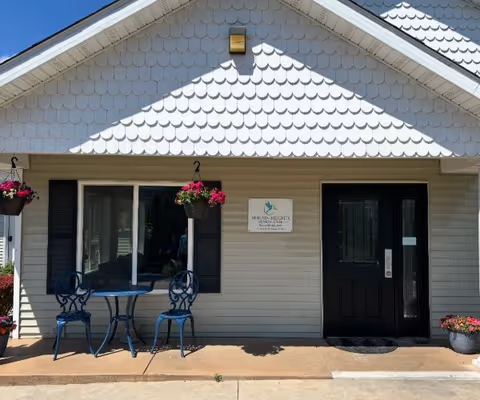 Front entrance of Auburn Heights Senior Care facility with a black door and a window with black shutters. There is a small blue metal table with two matching chairs on the porch, along with hanging flower baskets and a potted plant. The building exterior is light-colored with decorative shingles on the upper part of the wall.