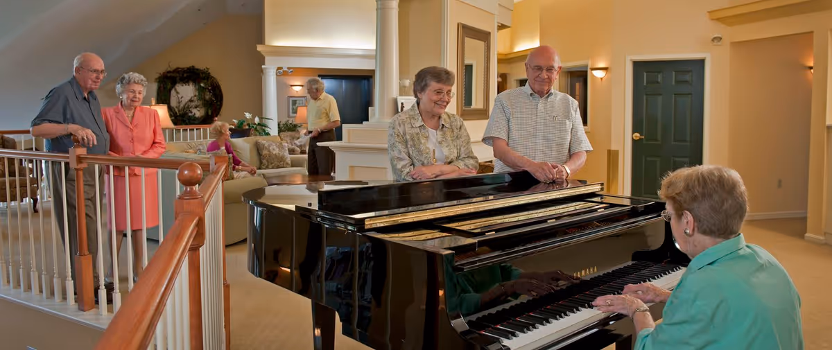 A group of elderly people gathered in a spacious, warmly lit common area. One woman is playing a black grand piano while others stand or sit nearby, smiling and enjoying the music. The room features beige walls, a green door, and comfortable seating in the background.