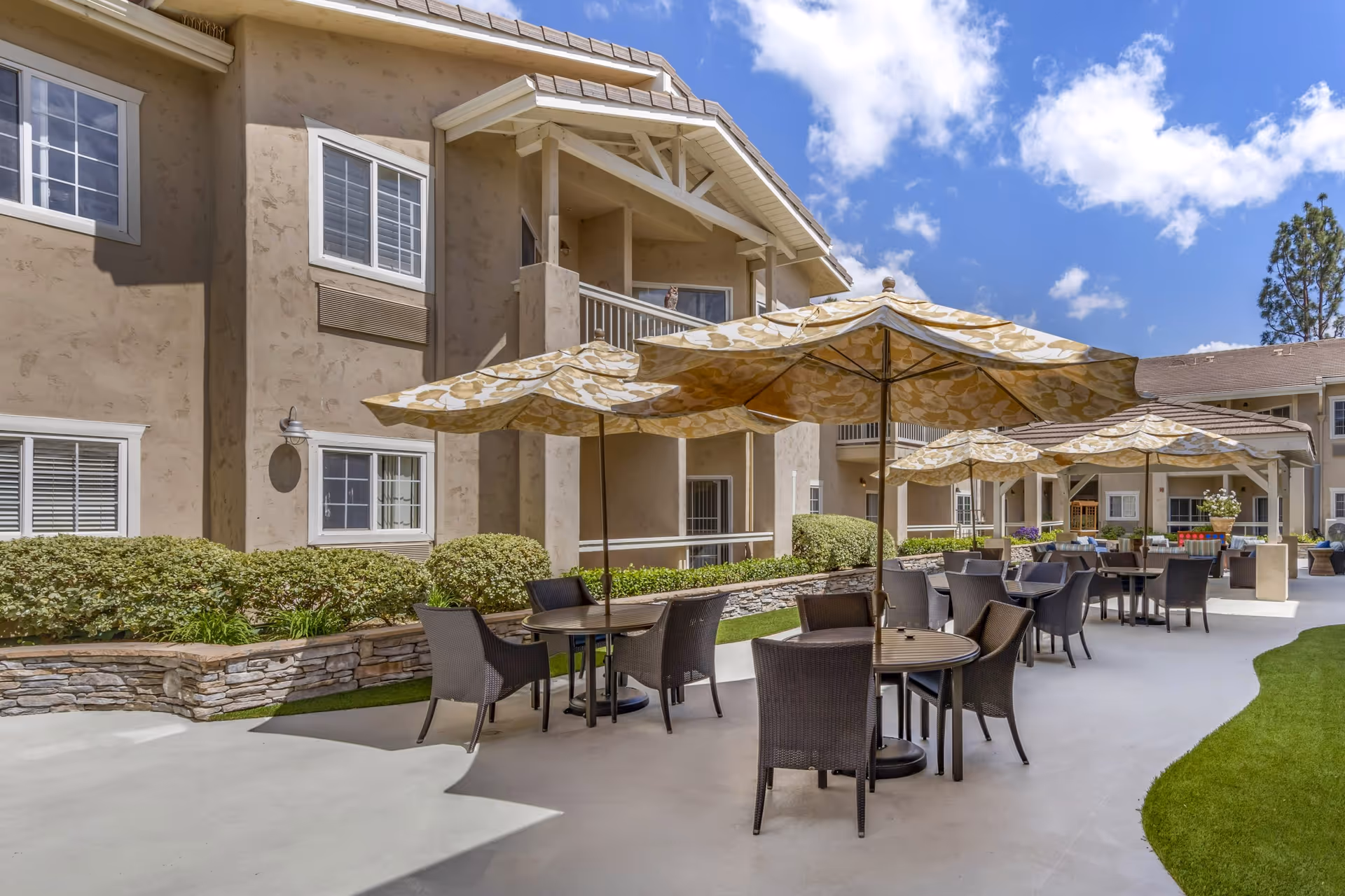 Outdoor patio area at Meadowbrook At Agoura Hills with round tables and wicker chairs under large patterned umbrellas, adjacent to a beige stucco building with windows and greenery, under a partly cloudy blue sky.