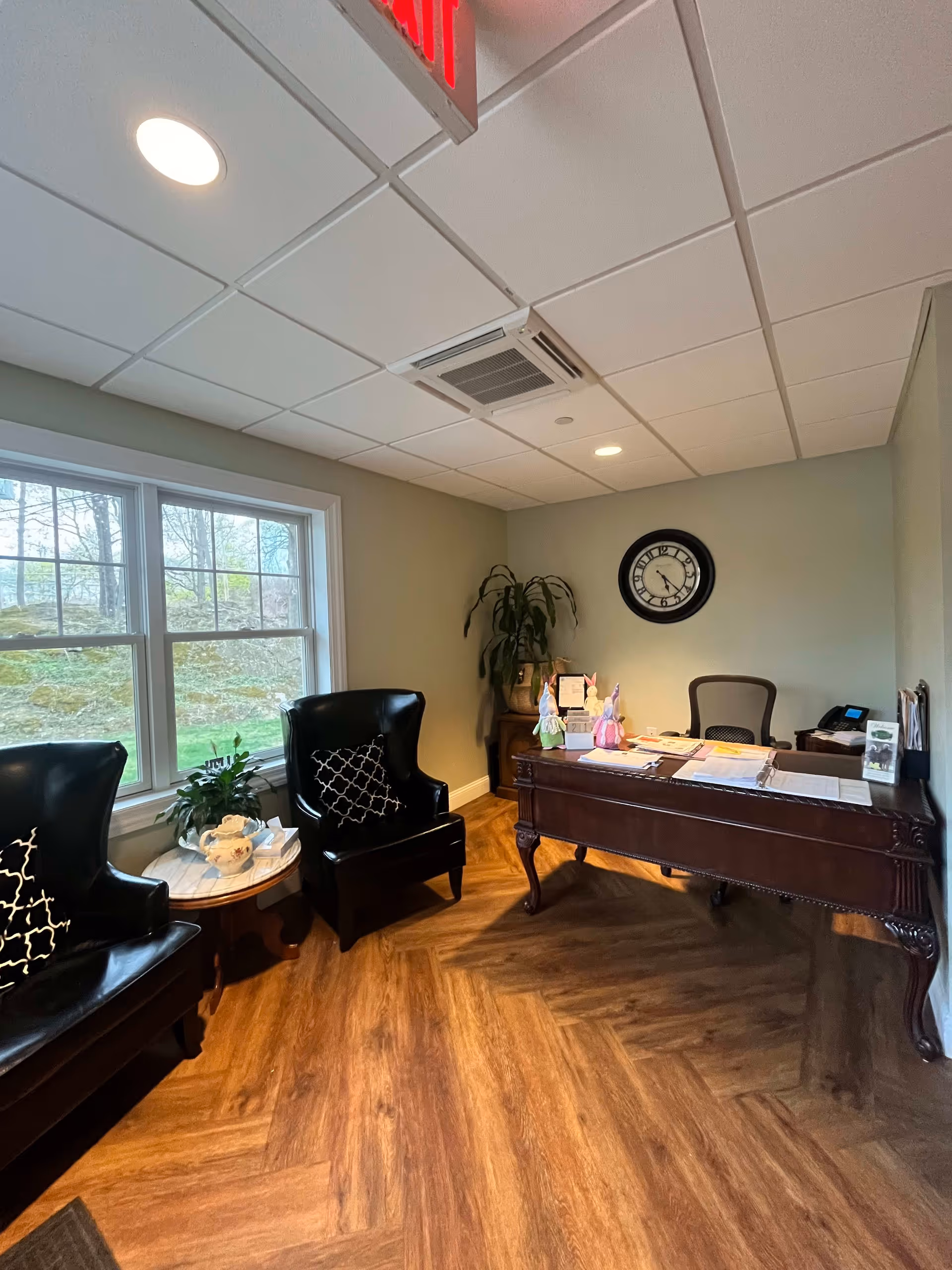 An office area with a wooden desk covered with papers and decorative items, a black office chair behind the desk, two black leather armchairs with patterned cushions, a small round table with a teapot and plant, a large window showing an outdoor view, a wall clock, and a potted plant in the corner.