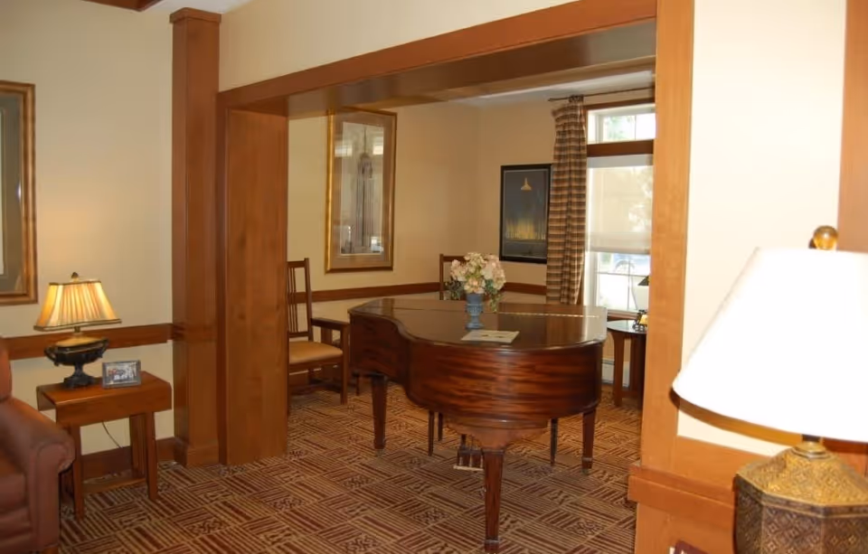 Interior view of a room with a wooden baby grand piano in the center, a vase with flowers on top, a wooden chair, framed artwork on the walls, a window with striped curtains, and a table lamp on a side table.