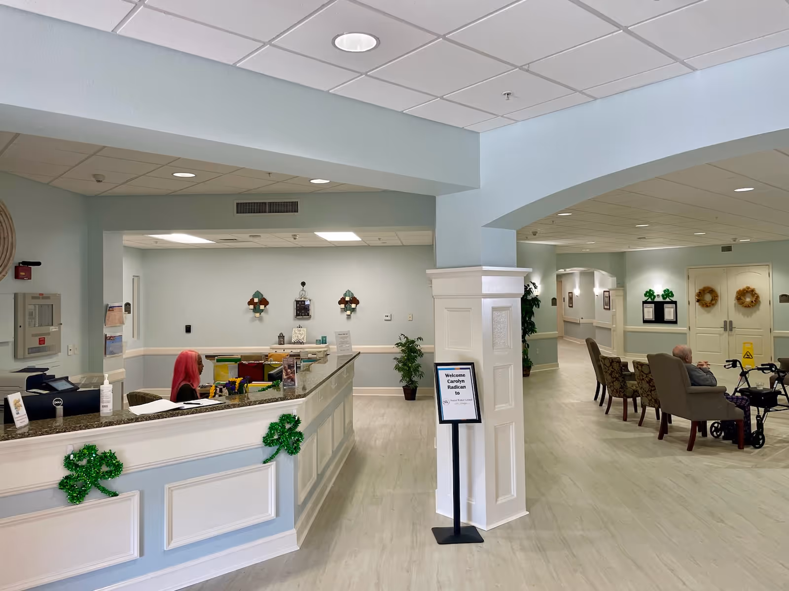 Interior view of a senior living facility reception area with a front desk where a person with pink hair is seated. The space has light blue walls, light wood flooring, and ceiling lights. There are green shamrock decorations on the front desk and a sign on a stand welcoming a visitor named Carolyn Radican. In the background, there is a seating area with chairs and an elderly person sitting with a walker nearby. The hallway extends further into the facility with doors and framed pictures on the walls.