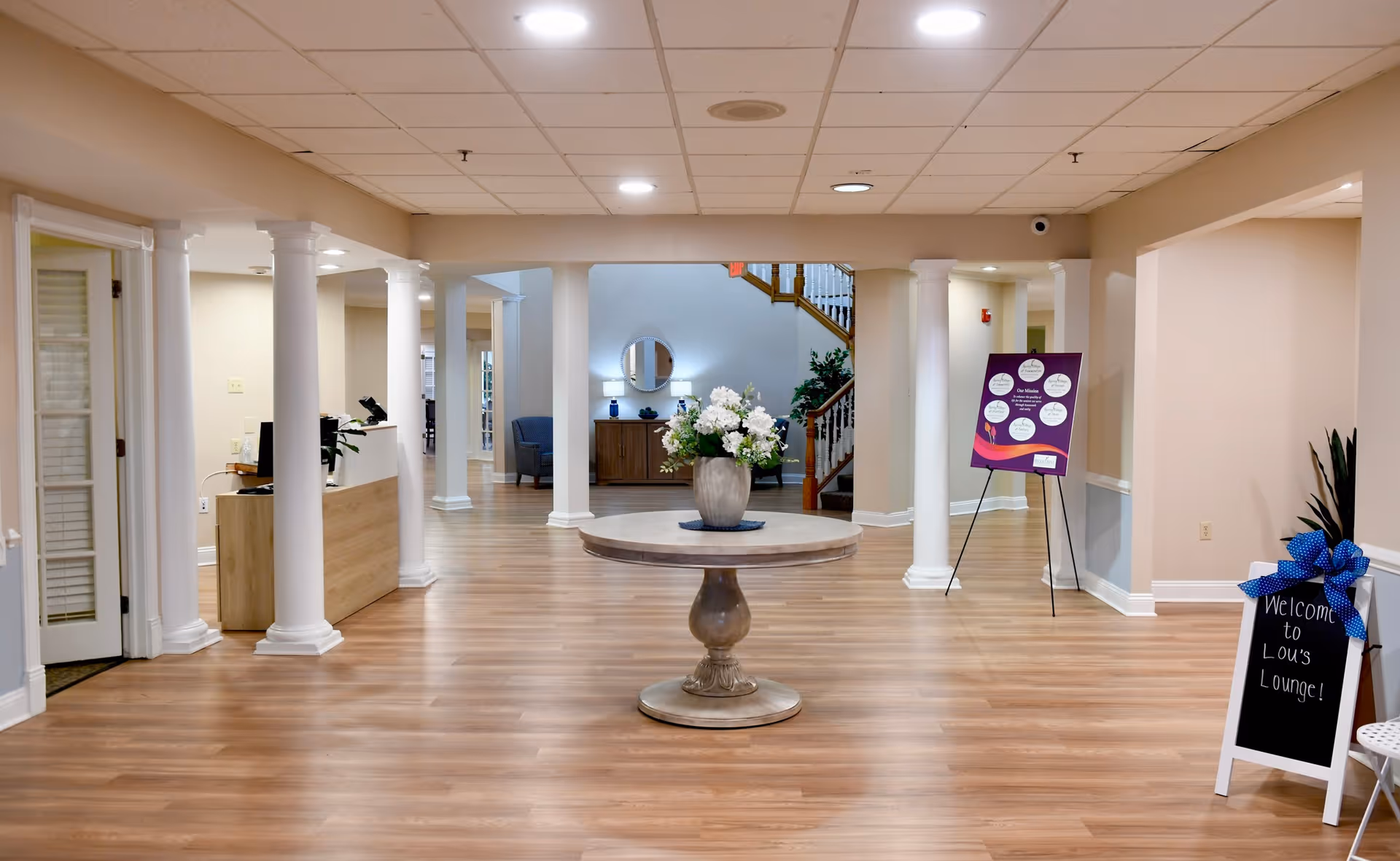 Spacious and well-lit interior of a senior living facility with wooden flooring, white columns, a round table with a flower arrangement in the center, a reception desk on the left, and a sign welcoming visitors to Lou's Lounge on the right.