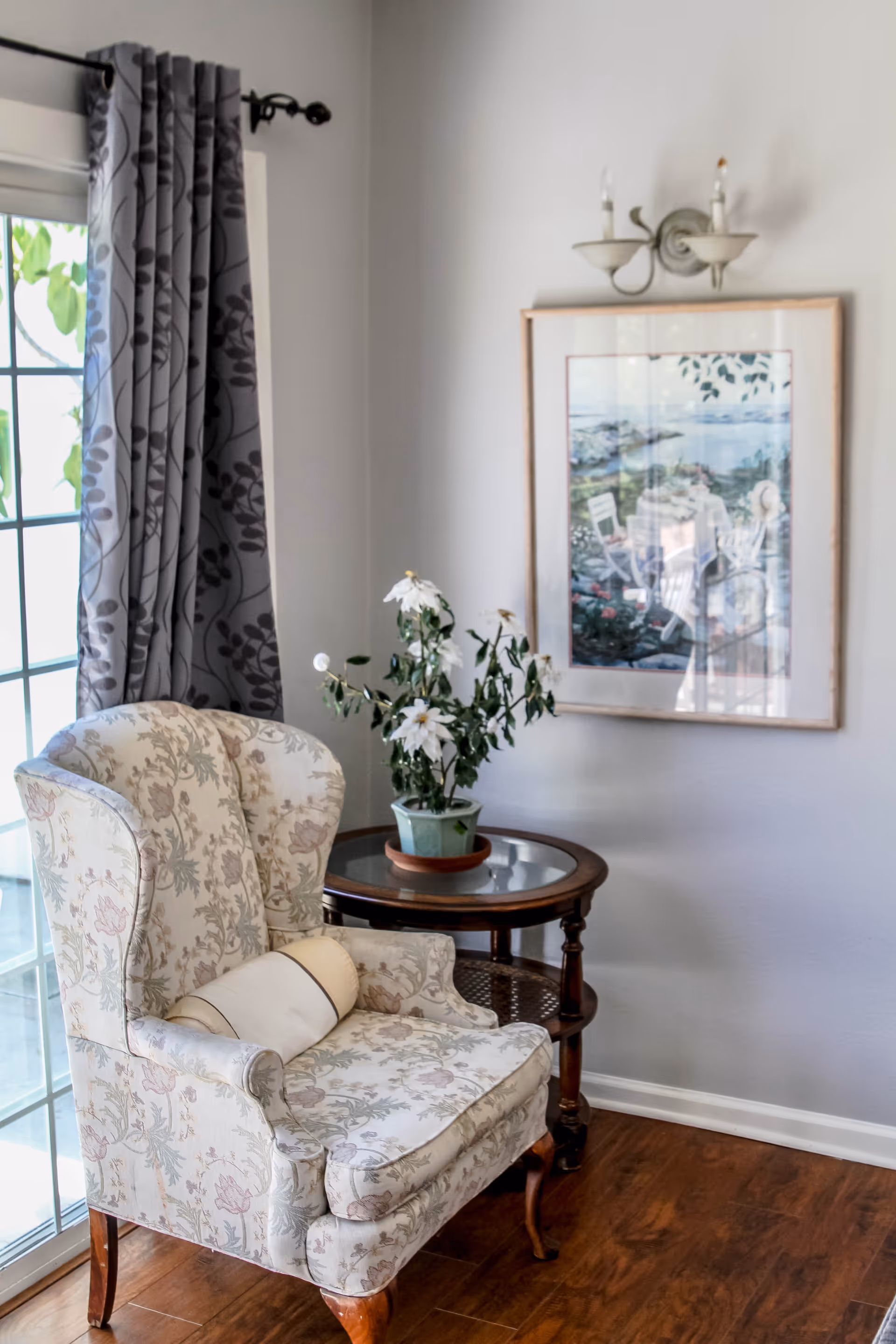 A cozy corner of a living room featuring a floral upholstered armchair with a small cylindrical pillow, next to a round wooden side table with a glass top holding a potted plant with white flowers. Behind the chair is a large window with patterned gray curtains, and on the wall hangs a framed painting of an outdoor scene with chairs and a table near water. The floor is wooden, and a two-light wall sconce is mounted above the painting.