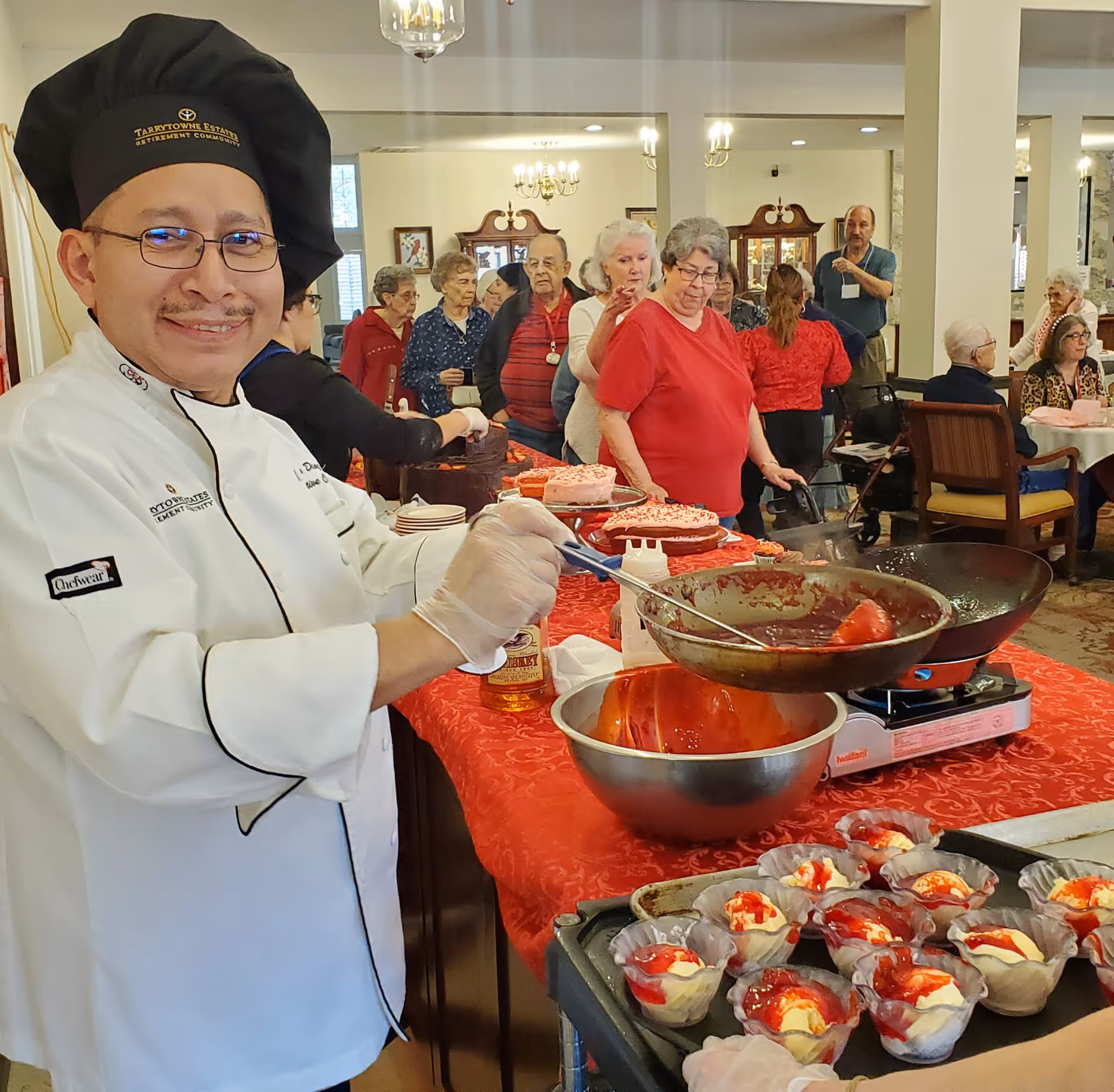 A smiling chef at Tarrytowne Estates serving red-topped desserts to residents gathered at a buffet in a dining/activity room.