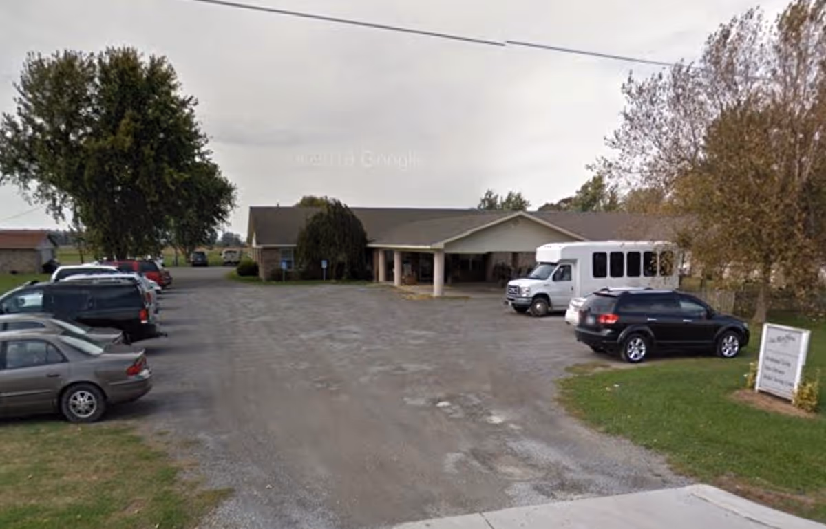 Single-story senior living building with a covered entrance, a gravel parking lot, several cars and a white passenger van.
