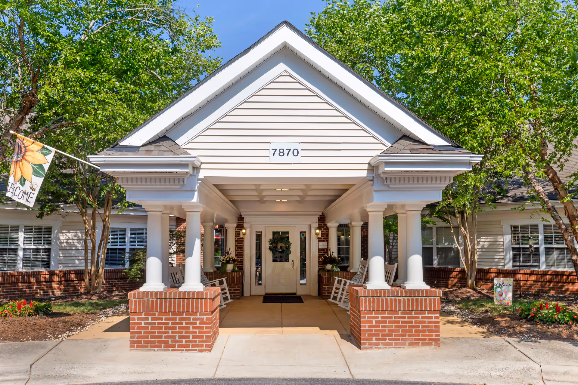 Front entrance of a senior living facility with a covered porch supported by white columns on brick bases, rocking chairs on either side, a door decorated with a wreath, and a flag with a sunflower and the word 'Welcome'. The building number 7870 is displayed above the entrance.