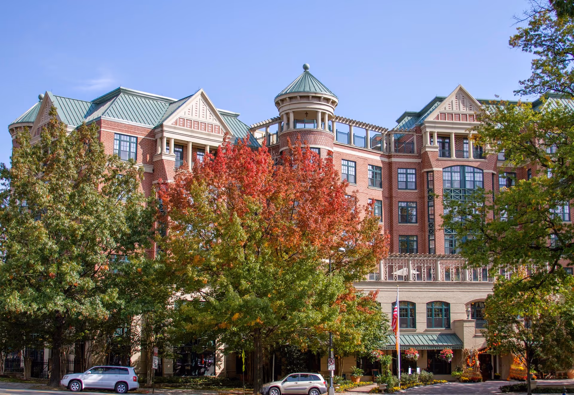 A large multi-story brick building with green roofs partially obscured by trees with green and red autumn leaves. There are parked cars in front of the building and a clear blue sky above.