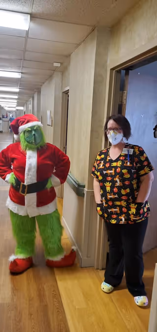 A person dressed as the Grinch in a Santa Claus outfit stands in a hallway next to a healthcare worker wearing a colorful scrub top with cartoon characters and a face mask. The hallway has beige walls, wooden flooring, and fluorescent ceiling lights.