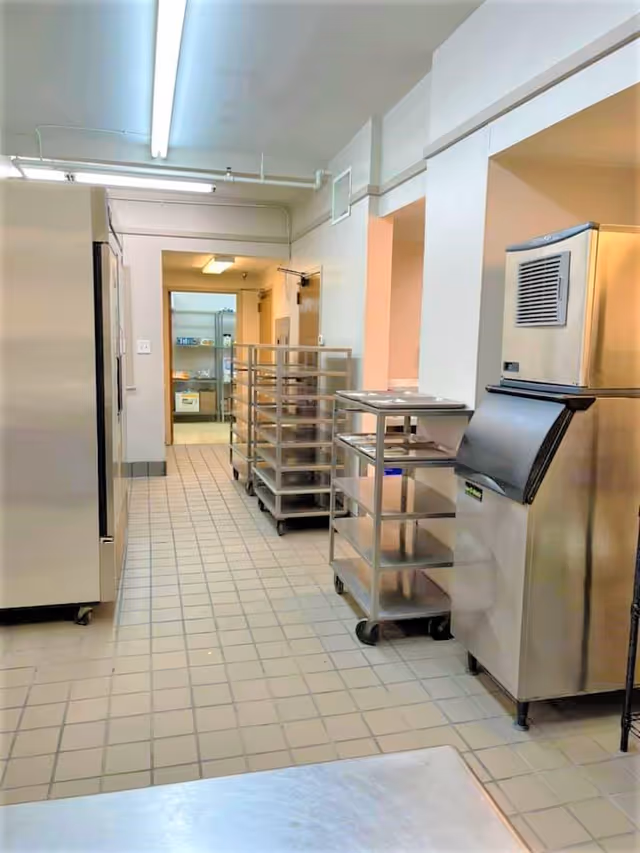 A clean commercial kitchen area with tiled floors, stainless steel appliances including a large refrigerator and an ice machine, and multiple metal racks on wheels for holding trays or food items. The space is well-lit with fluorescent lighting and has a doorway leading to another room with more shelving and supplies.