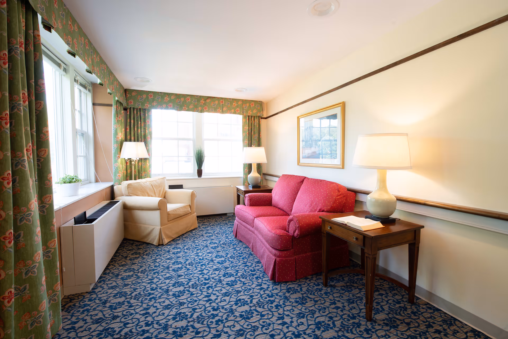 A cozy sitting room with a red loveseat, beige armchair, wooden side tables with lamps, patterned curtains and blue carpet.