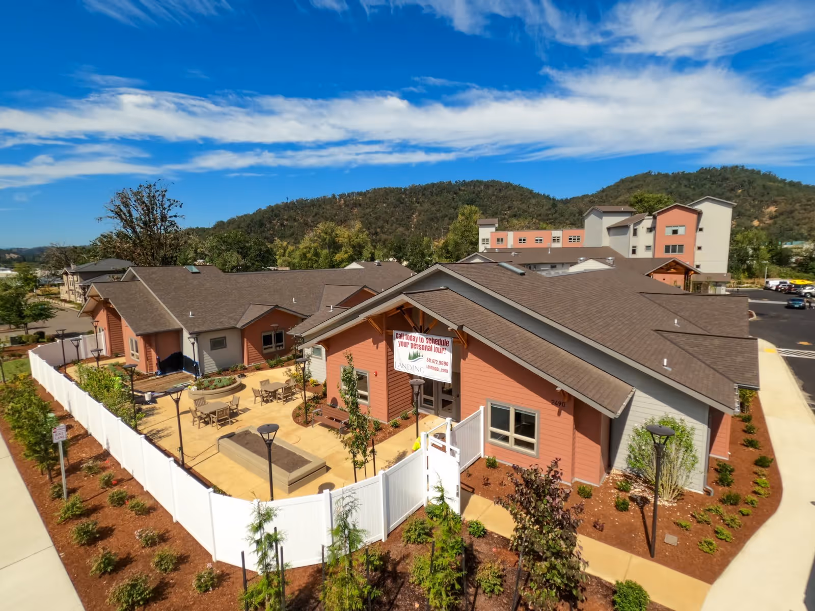 Aerial view of The Landing a Senior Living Community showing multiple connected buildings with brown roofs and peach-colored walls. There is a fenced outdoor patio area with tables and chairs, surrounded by landscaped plants and trees. Hills and a blue sky with scattered clouds are visible in the background.