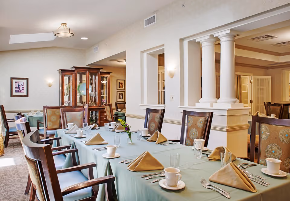 A well-lit dining room with a long table covered in a light green tablecloth, set with beige folded napkins, white cups and saucers, glasses, and silverware. The room features wooden chairs with patterned upholstery, a wooden china cabinet with glass doors displaying dishes, and decorative columns separating the dining area from an adjacent room.