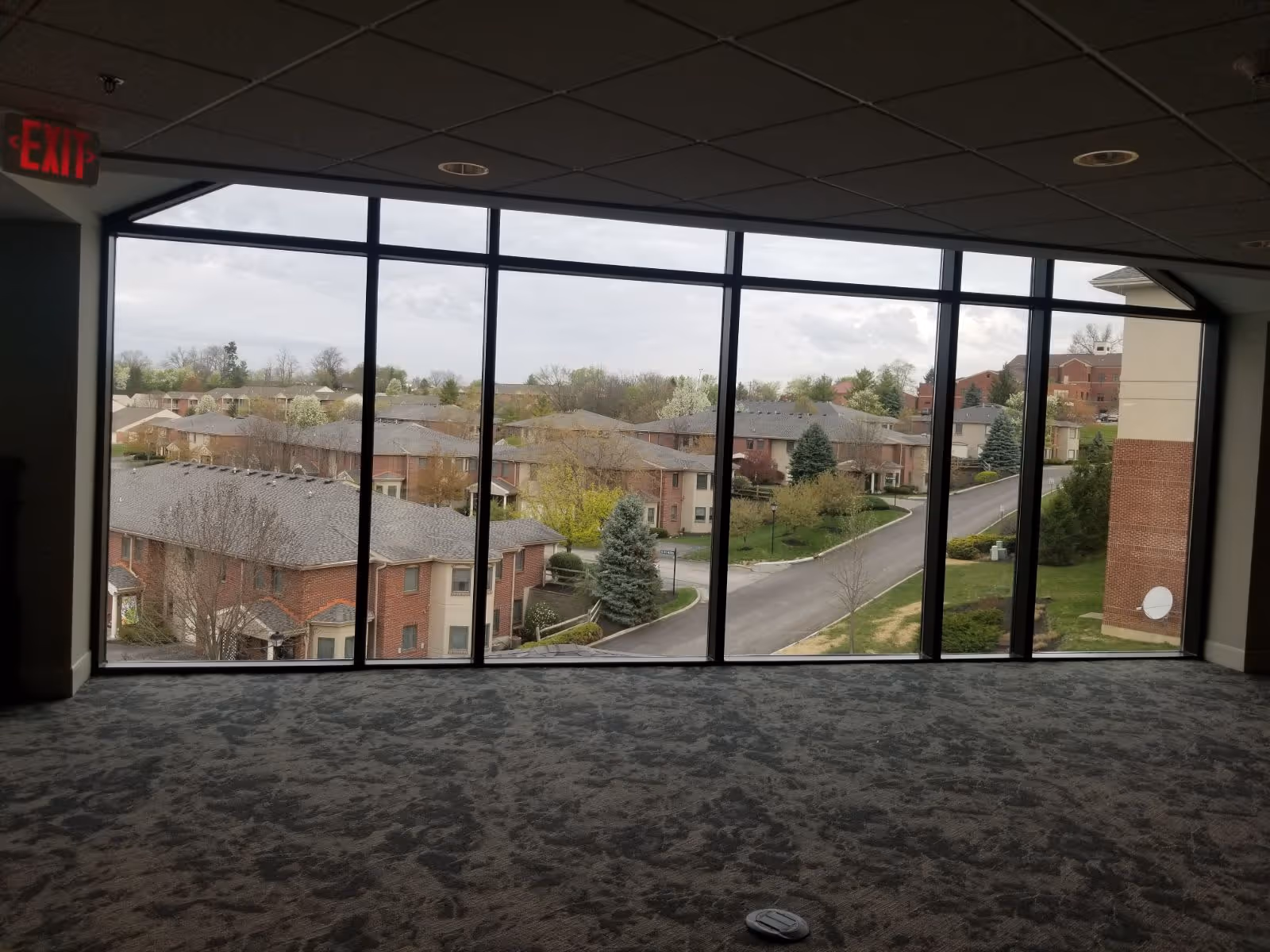 Carpeted interior room with large floor-to-ceiling windows overlooking residential buildings and a road.