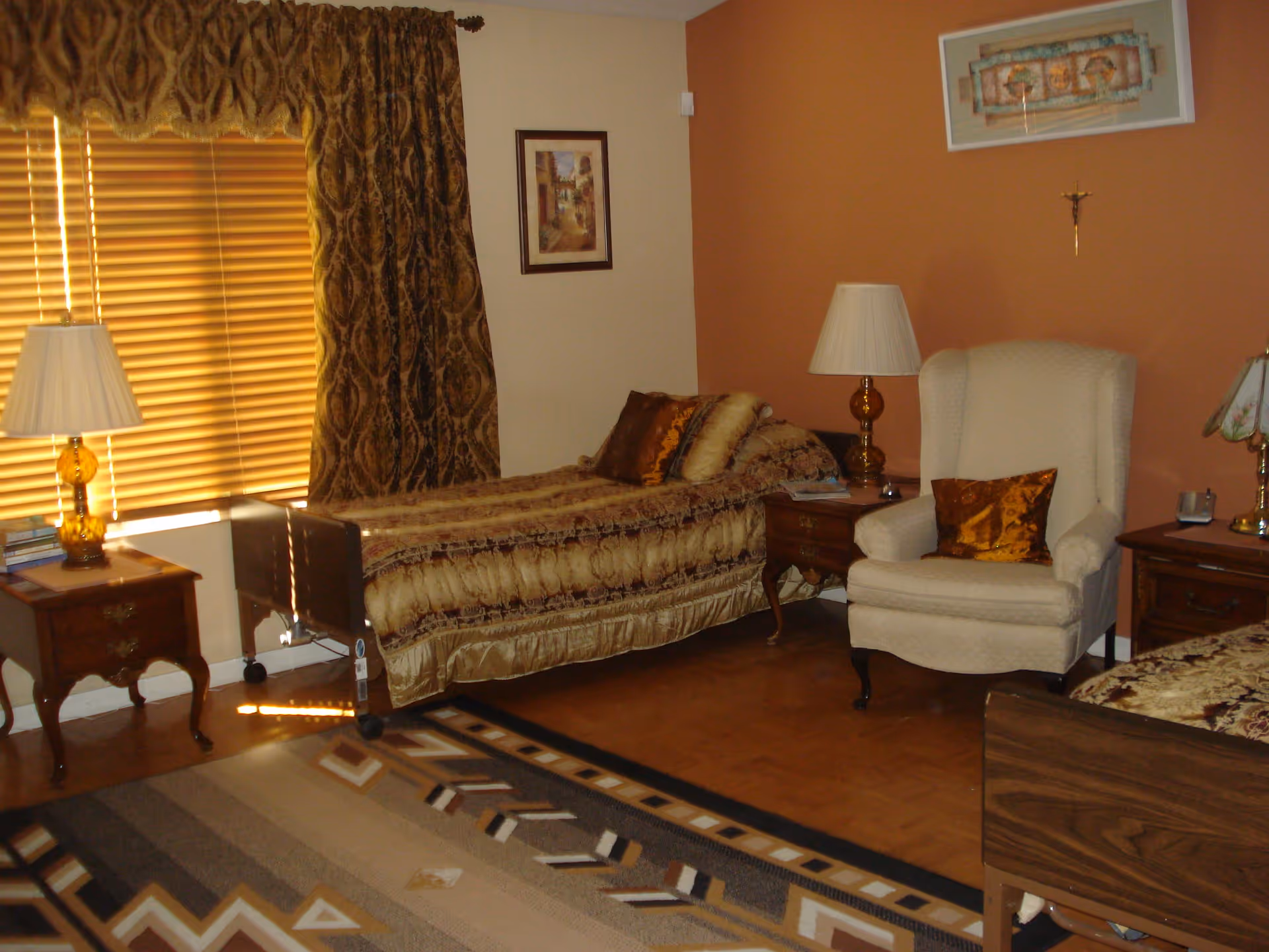 A cozy assisted living bedroom with two beds covered in patterned bedspreads, a beige armchair with a decorative pillow, two wooden nightstands with lamps, a window with closed blinds and heavy curtains, a framed picture on the wall, and a patterned area rug on the floor.
