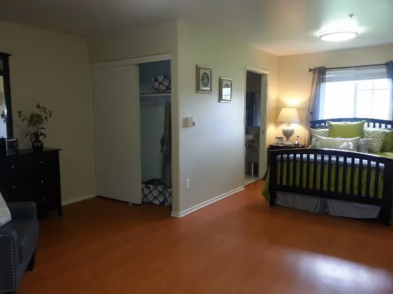 A bedroom with a wooden bed frame and green bedding, a nightstand with a lamp, a window with curtains, an open closet with sliding doors, a dresser with a vase of flowers, and a chair. The room has wooden flooring and light-colored walls.