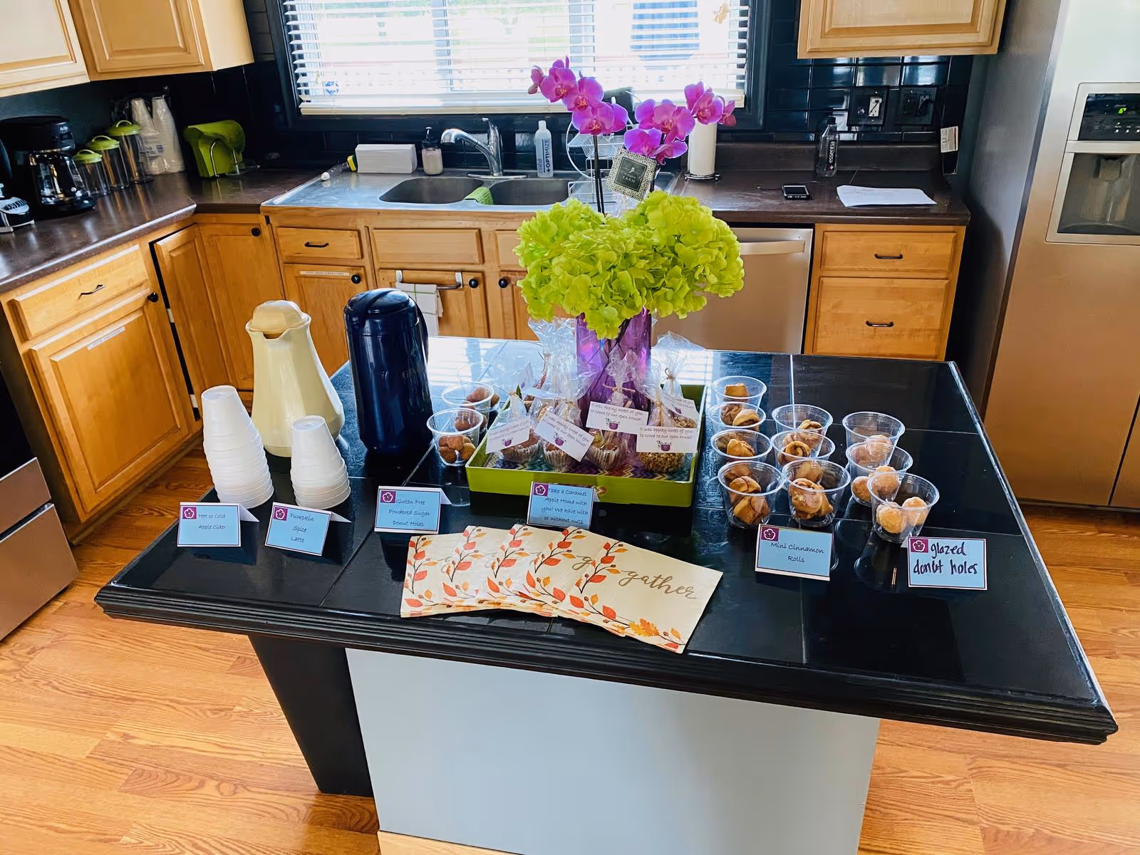 Kitchen island with coffee dispensers, disposable cups, labeled snack cups, and a floral centerpiece in a residential kitchen.