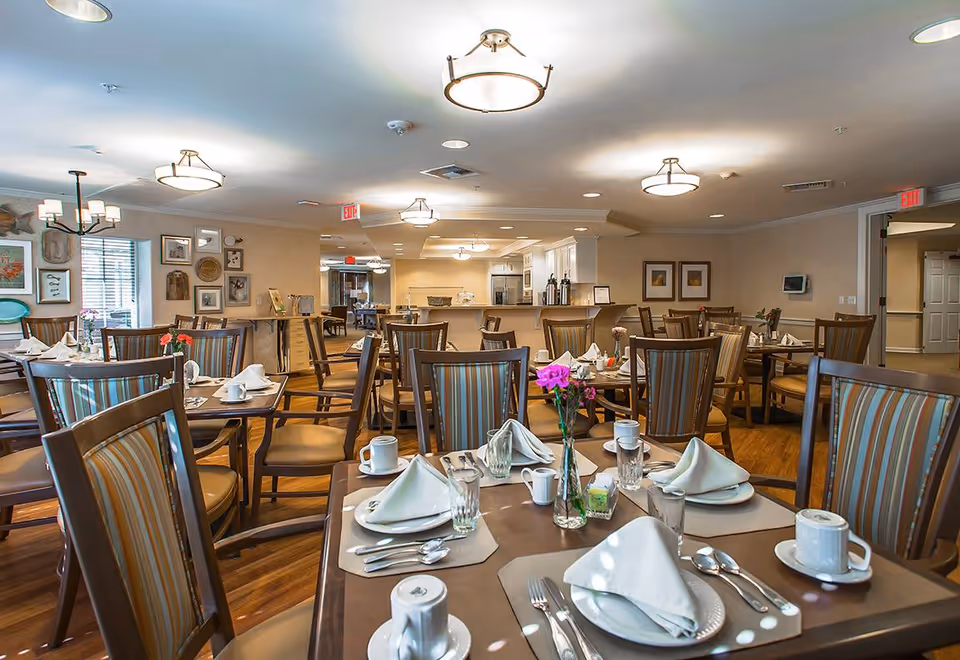 A well-lit dining room in a senior living facility with multiple tables set with white napkins, plates, cups, glasses, and silverware. The chairs have striped upholstery, and there are small flower vases on the tables. The room has wooden flooring, framed artwork on the walls, and ceiling lights. In the background, there is a kitchen area with white cabinets and coffee dispensers.