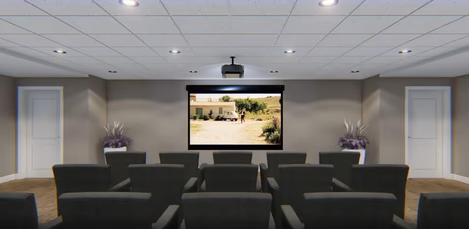 A small theater room with two rows of dark gray cushioned chairs facing a wall-mounted projector screen showing a scene from a movie. The room has beige walls, a white ceiling with recessed lighting, two white doors on either side of the screen, and two large white planters with purple plants placed symmetrically near the doors.