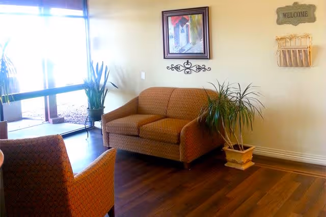 A cozy seating area with an orange patterned loveseat and armchair on a wooden floor. There are two potted plants, one on the floor and one on a small table near a glass door letting in natural light. The wall features a framed picture, a decorative metal wall piece, and a wooden 'WELCOME' sign with a small rack beneath it.