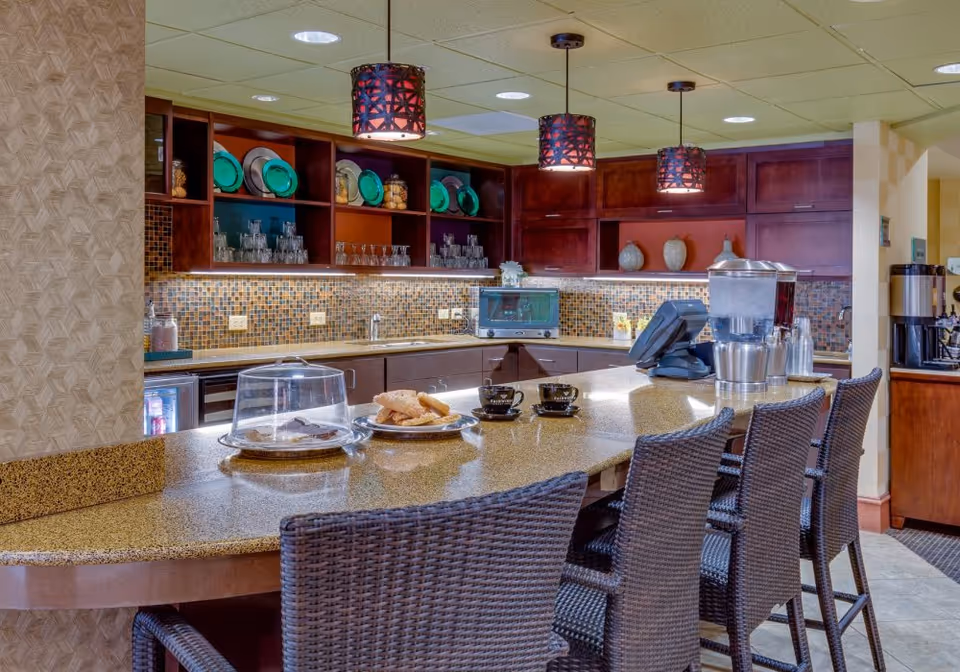 A communal kitchenette and breakfast bar with wicker barstools, pendant lights, a countertop with pastries and coffee equipment, and shelves of dishes and glassware.