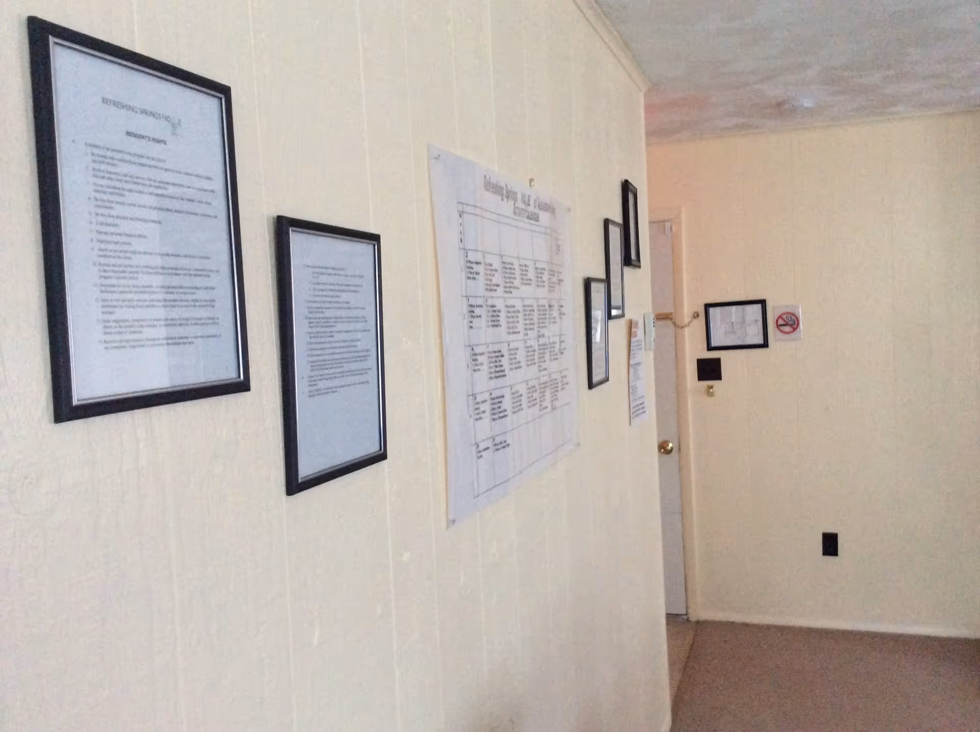 A beige hallway wall with multiple framed documents and notices hung in a row. The floor is carpeted, and there is a white door at the end of the hallway. The wall has a no smoking sign and a small framed floor plan.