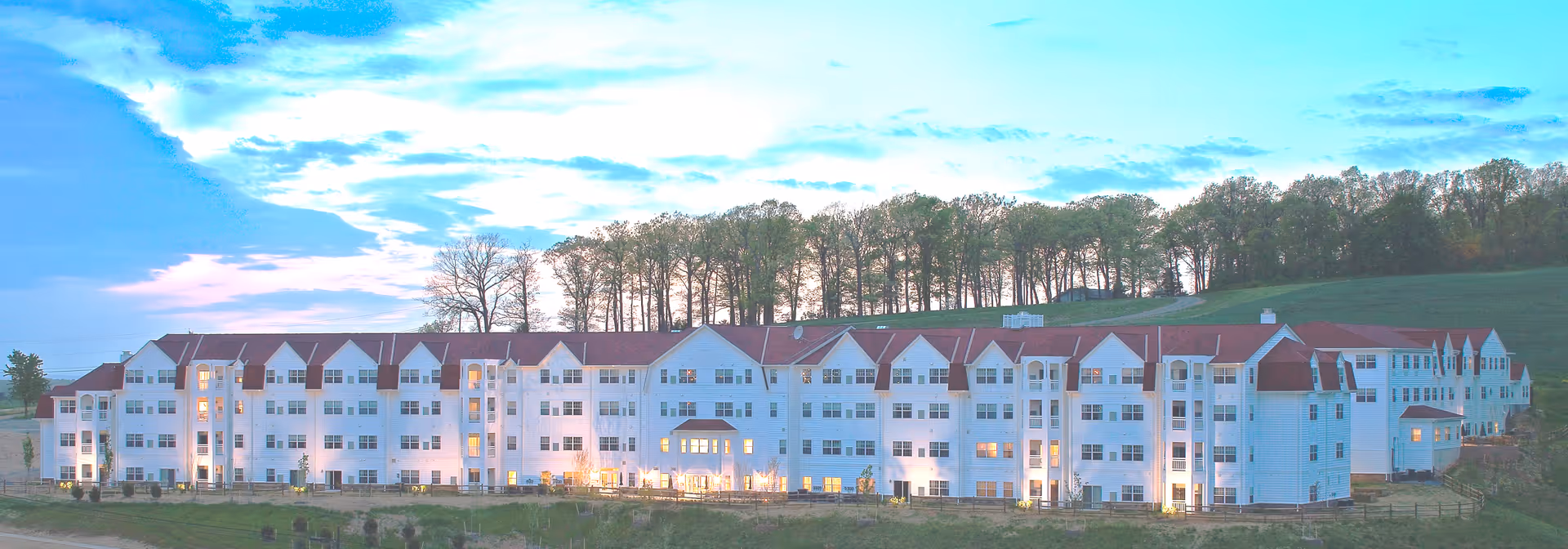 Wide exterior view of a large, multi-story senior living facility building with white walls and red roofs, surrounded by grass and trees under a partly cloudy sky at dusk.
