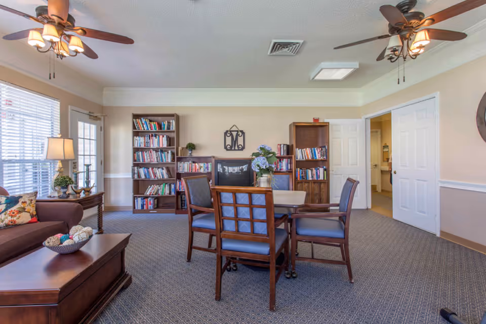 A cozy common area with a table and four chairs in the center, two wooden bookshelves filled with books against the back wall, a brown sofa with floral pillows on the left, a wooden coffee table with decorative balls, two ceiling fans with lights, and a door with windows letting in natural light.