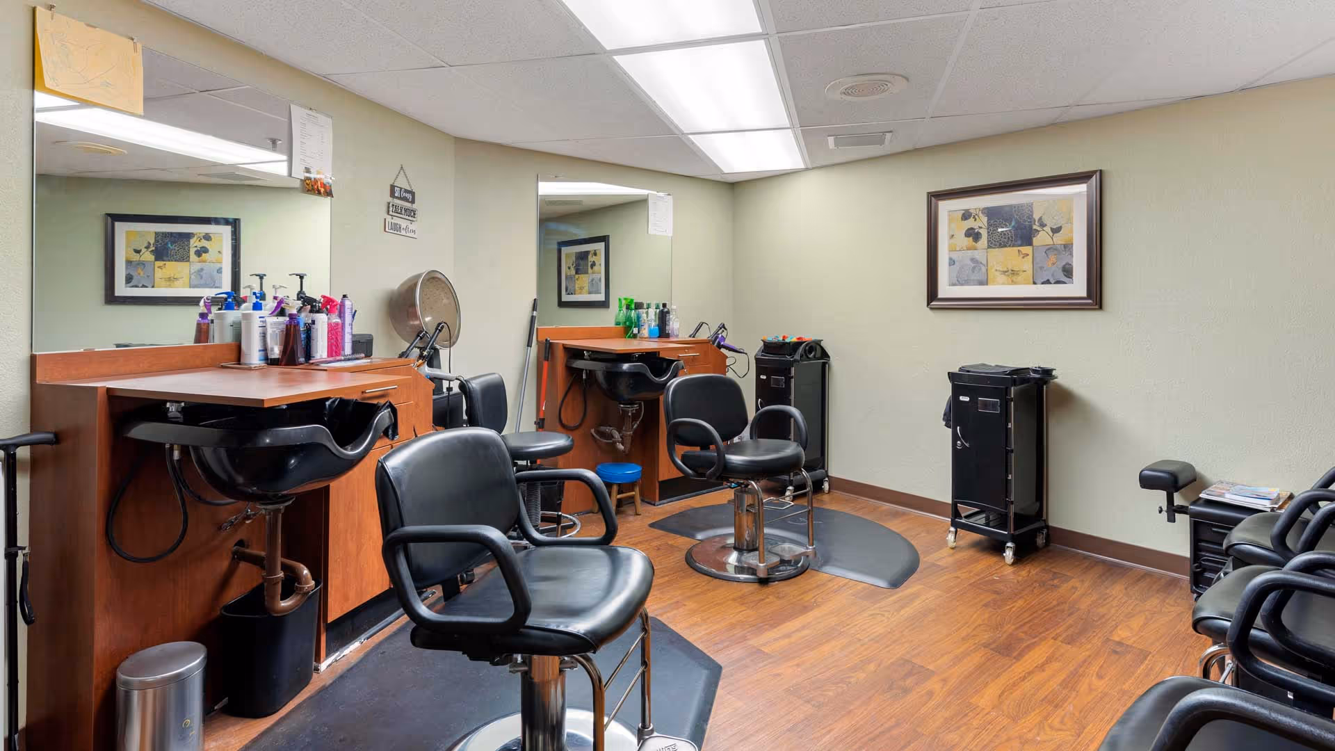 Interior view of a hair salon area with two black salon chairs in front of wooden counters with sinks and various hair care products. The room has light green walls, a framed artwork, and wooden flooring.