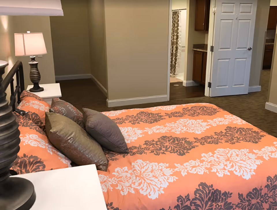 A bedroom with a bed covered in an orange, white, and brown patterned comforter and several pillows. There are two bedside tables with matching lamps. In the background, an open door reveals a bathroom with a shower curtain and a glimpse of a kitchen area.