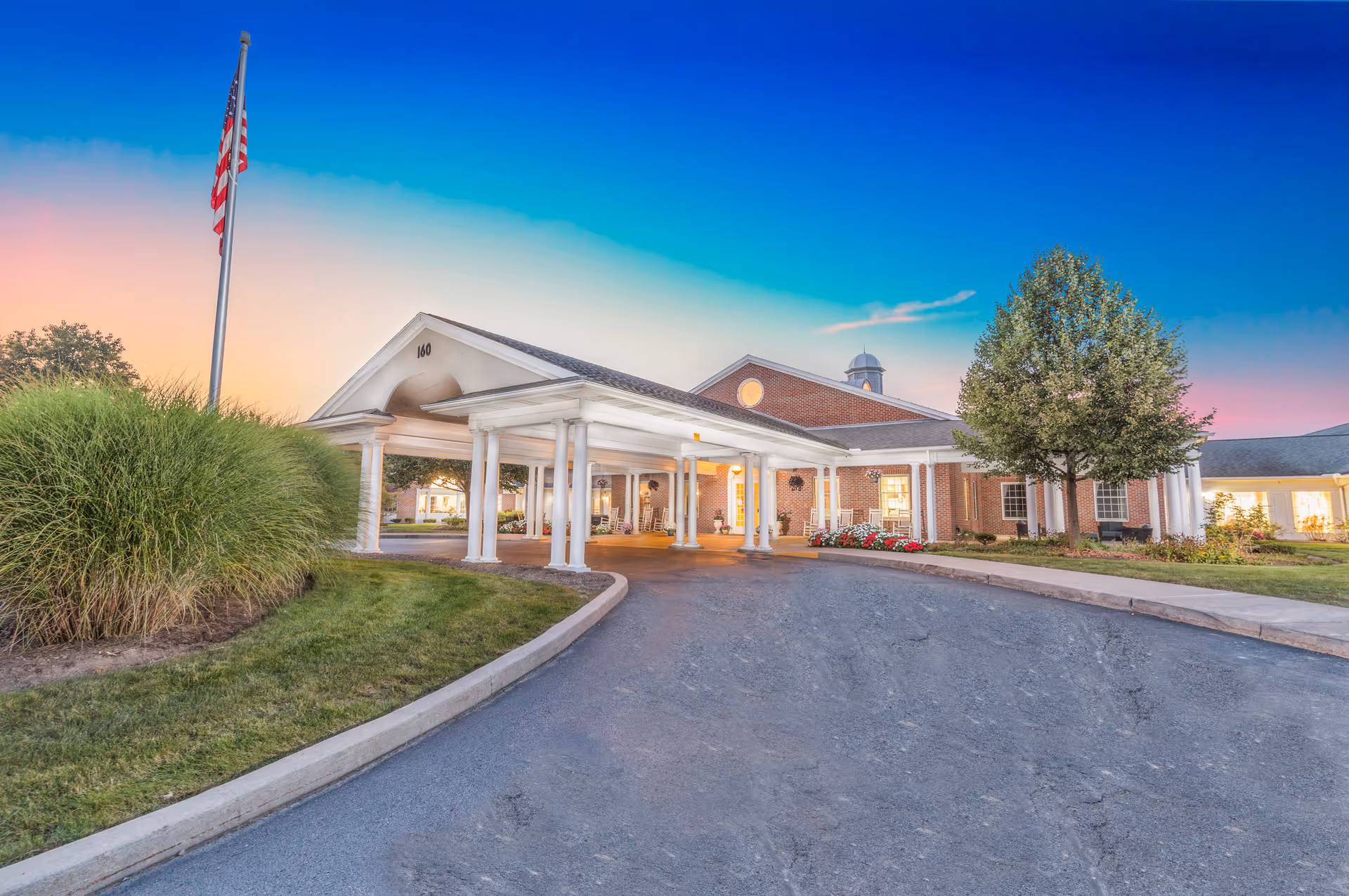 Exterior view of Heather Heights: Assisted Living & Memory Care Pittsford building at sunset, showing a covered entrance with white columns, a driveway, an American flag on a flagpole, manicured grass, and trees.