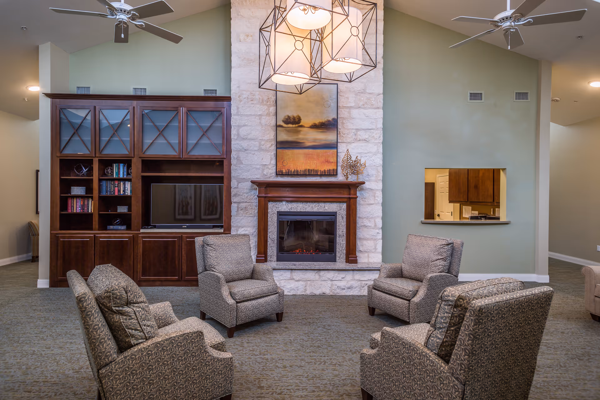 A cozy living room area with four patterned armchairs arranged around a stone fireplace with a wooden mantle. Above the fireplace hangs a landscape painting. To the left is a dark wooden entertainment center with shelves, books, and a TV. The walls are painted light green, and there are ceiling fans and modern pendant lights hanging from the ceiling. A small pass-through window to a kitchen area is visible on the right wall.