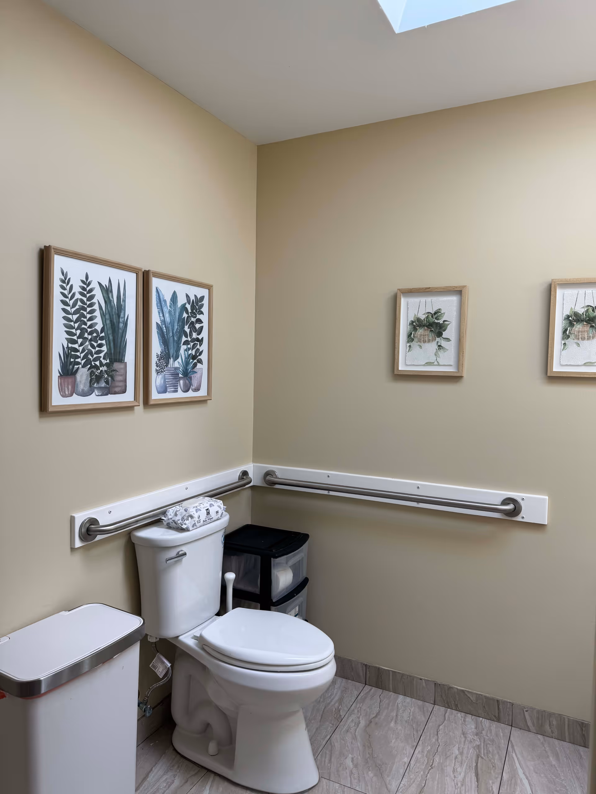 A clean bathroom with beige walls and tiled floor. There is a white toilet with a folded towel on top of the tank, a white trash bin with a silver lid, and a small black storage unit with drawers next to the toilet. Two metal grab bars are mounted on the walls around the toilet. Four framed botanical prints hang on the walls. A skylight is visible on the ceiling.