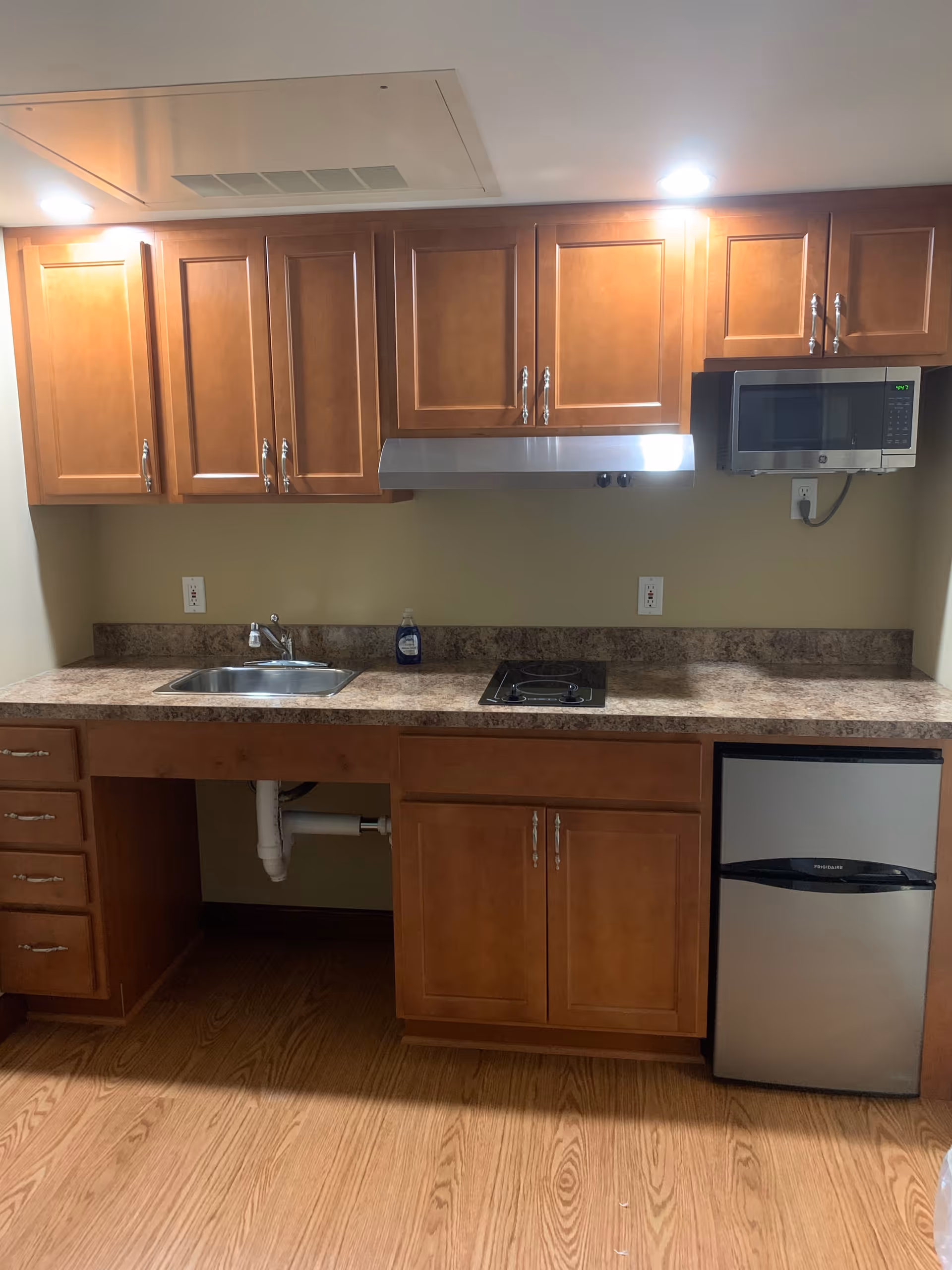 A small kitchen area with wooden cabinets, a stainless steel sink, a two-burner electric cooktop, a stainless steel microwave mounted above the cooktop, and a compact stainless steel refrigerator. The countertop is a brown marble pattern, and the floor is wood.