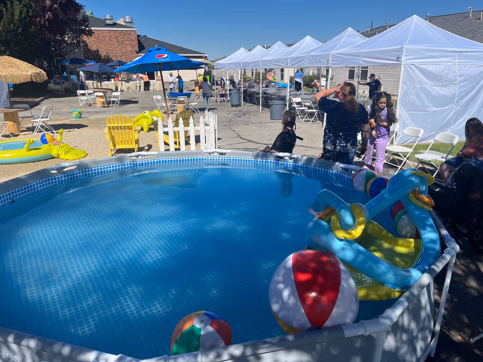 Outdoor area at Country Pines Retirement Community featuring an above-ground pool with colorful inflatable toys and beach balls. Several people are gathered around under white canopy tents and blue umbrellas, with folding chairs and tables set up on a sunny day.