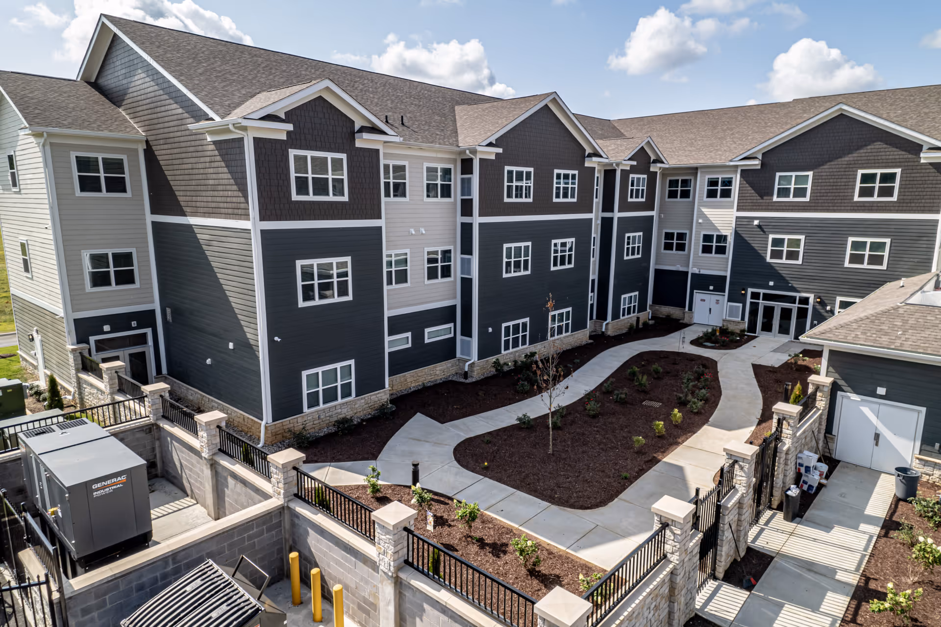 Exterior view of a modern multi-story senior living facility building with gray and white siding, multiple windows, and a landscaped courtyard with pathways and small plants under a partly cloudy sky.