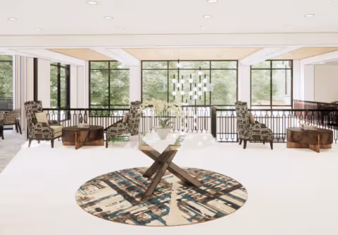 A bright and spacious living room area with large windows showing greenery outside. The room features four patterned armchairs arranged around two wooden side tables, and a glass coffee table with a vase of white flowers on a round patterned rug. Modern pendant lights hang from the ceiling.