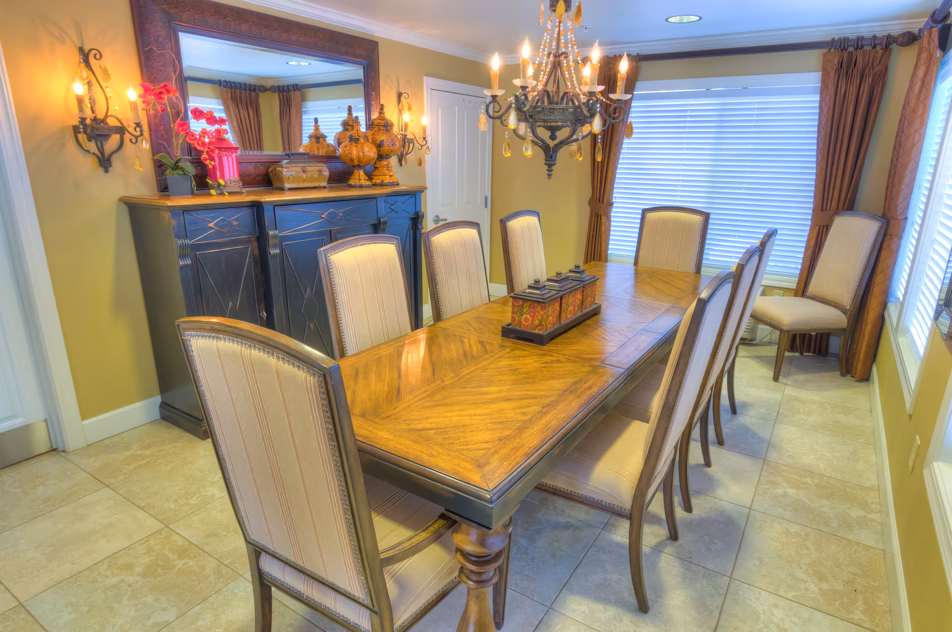 A well-lit dining room with a long wooden dining table surrounded by eight upholstered chairs. The room features a large mirror above a dark wooden sideboard decorated with vases and a pink orchid. A chandelier hangs above the table, and large windows with brown curtains allow natural light to fill the space.