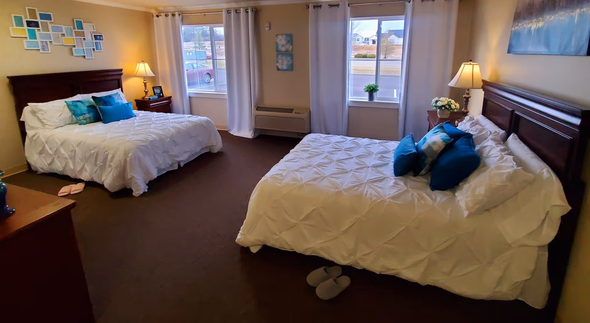 A bedroom with two beds featuring white comforters and blue accent pillows. Each bed has a wooden headboard and a nightstand with a lamp. There are two windows with white curtains letting in natural light, and slippers are placed on the carpeted floor near each bed. The walls are decorated with framed artwork.