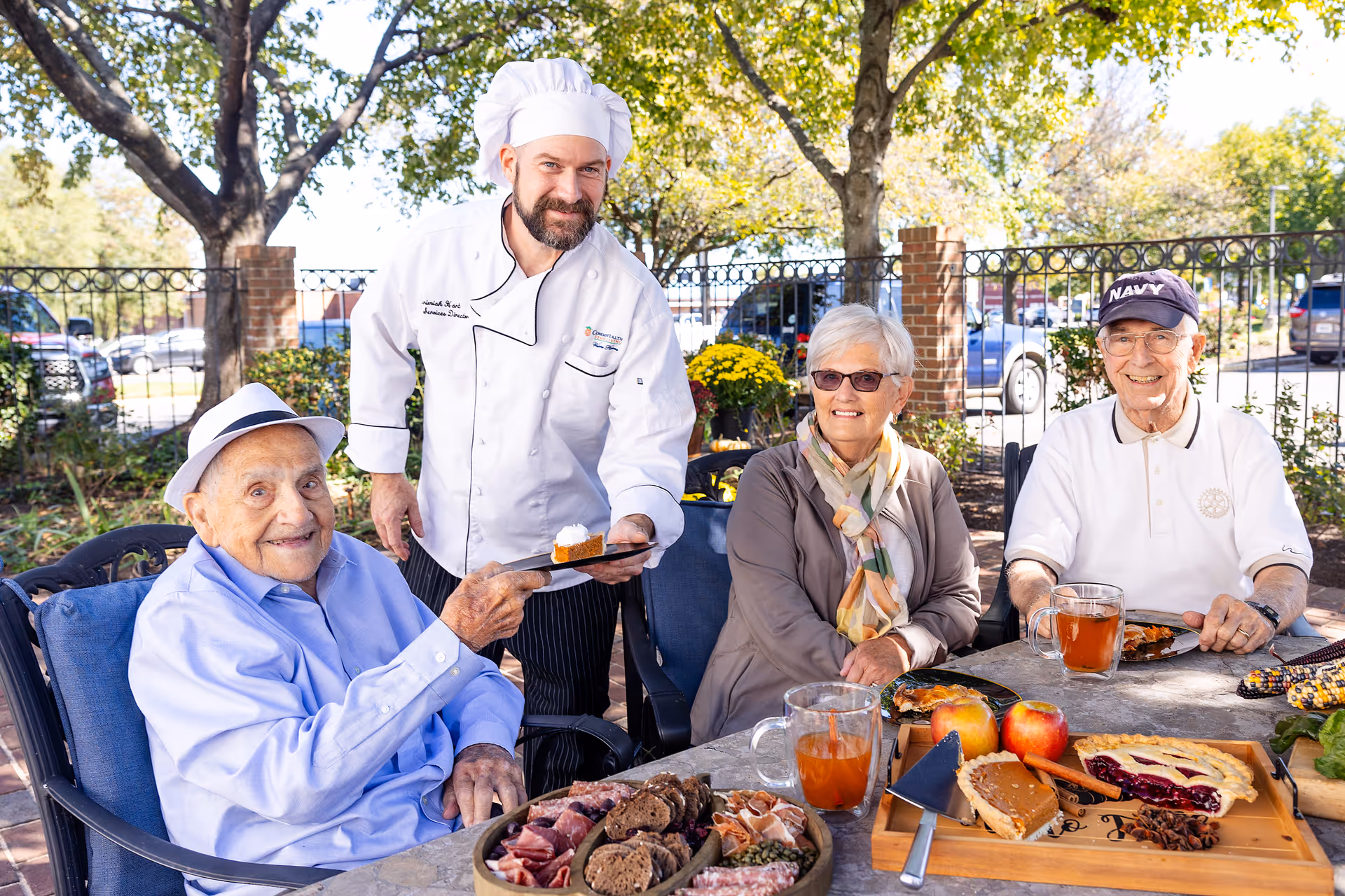 A chef in a white uniform and hat serves a slice of pie to an elderly man wearing a light blue shirt and white hat, seated at an outdoor table. Two other elderly individuals, a woman with short white hair and sunglasses and a man wearing a navy cap and white polo shirt, sit at the table with plates of food and drinks. The setting is a sunny outdoor patio with trees and a black metal fence in the background.