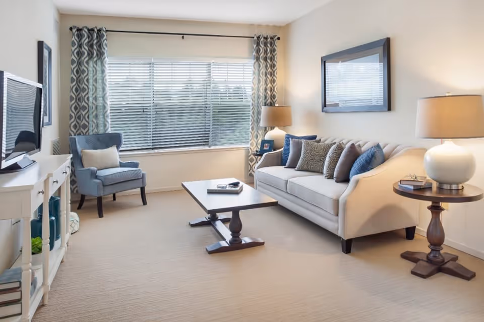 A bright living room with a large window covered by blinds and patterned curtains. The room features a beige sofa with multiple decorative pillows, a wooden coffee table, a blue upholstered armchair with a white cushion, two side tables with lamps, and a flat-screen TV on a white console table. The walls are light-colored, and the floor is carpeted.