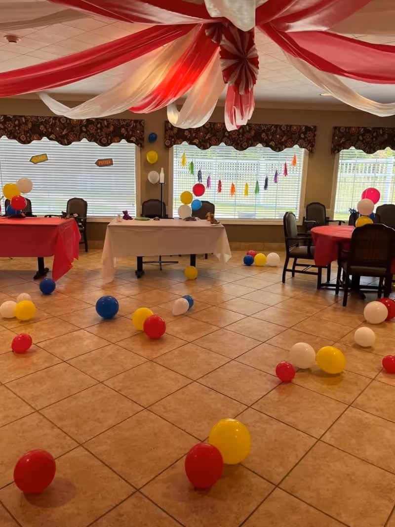 A decorated room with red, white, yellow, and blue balloons scattered on the tiled floor and tables covered with red and white tablecloths. Red and white fabric drapes hang from the ceiling, and colorful tassel garlands are displayed on the windows. Several chairs are arranged around the tables.