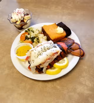 A plate of food featuring a lobster tail with lemon slices, steamed vegetables including broccoli, cauliflower, and carrots, a serving of cornbread with butter, and a small cup of melted butter. Next to the plate is a dessert cup with whipped cream and chocolate sauce.