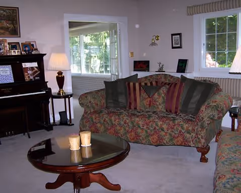 Cozy living room with a floral-upholstered sofa, round glass-top coffee table with candles, piano, and lamps near windows.